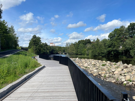 Ett promenaddäck i trä med svart staket vid ett vattendrag.