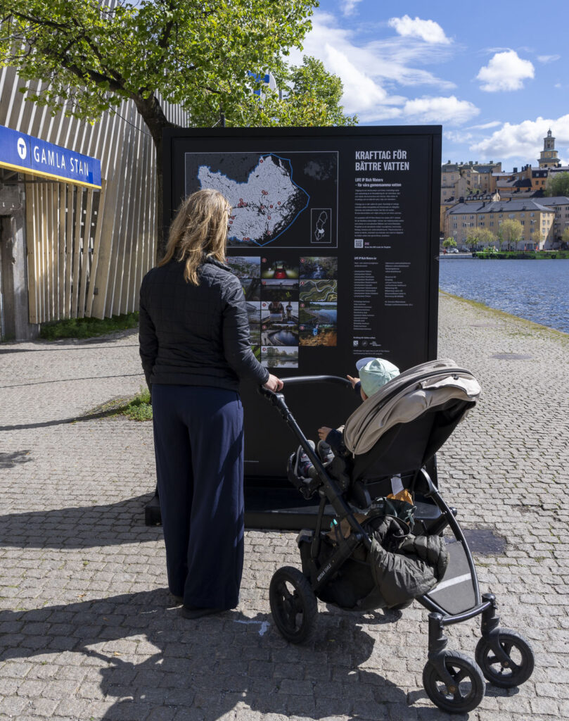 En kvinna med barnvagn läser på en stor skylt med en karta. Tunnelbanenedgång med skylten Gamla stan syns till vänster.