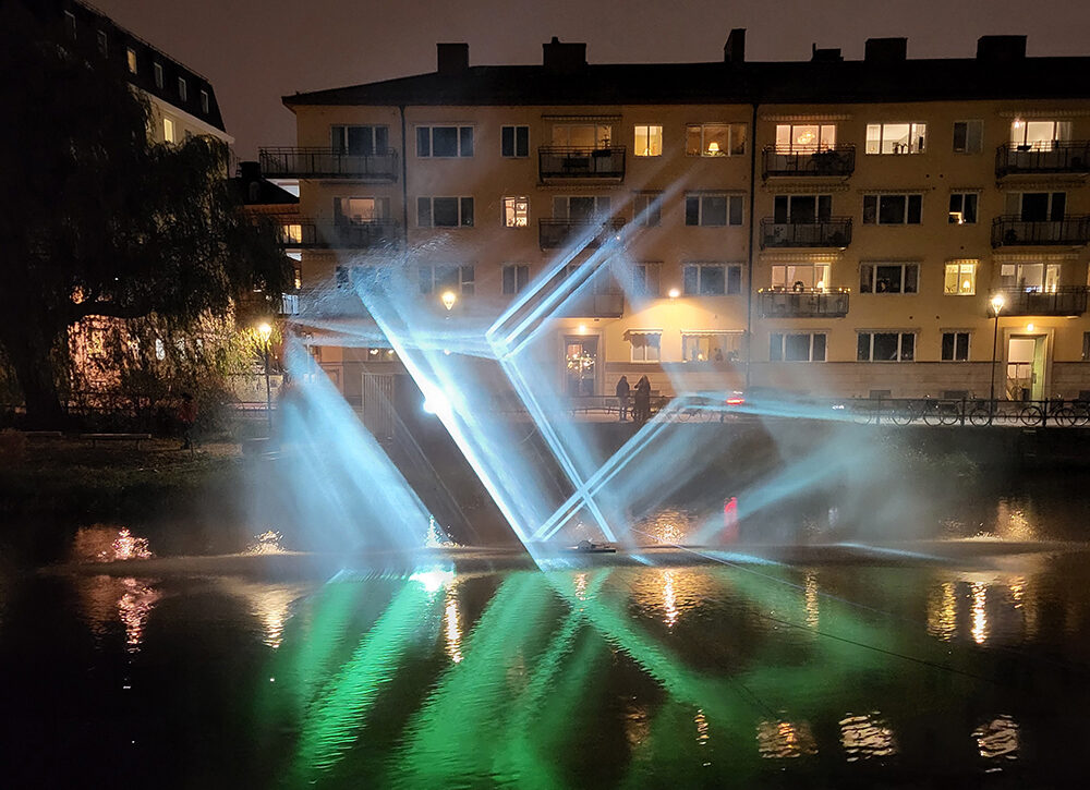 Ett blått och grönt ljussken i abstrakta former över vatten. I bakgrunden syns stadsbebyggelse.