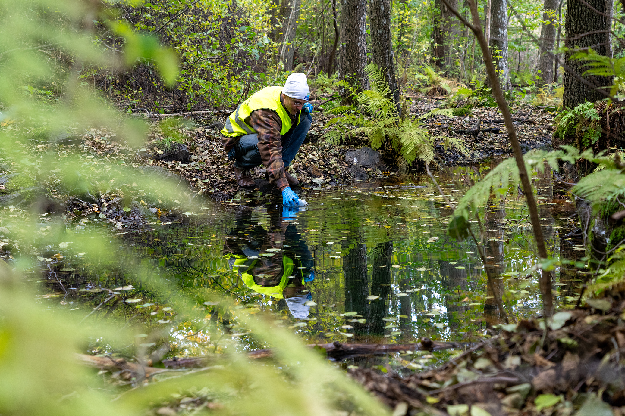 En person sitter på huk vid ett stilla vatten i en skog och håller ner ett mätinstrument i vattnet.