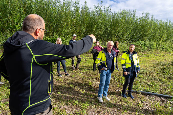 En grupp personer står och tittar på en man i svart jacka som pekar mot en plantering med höga växter